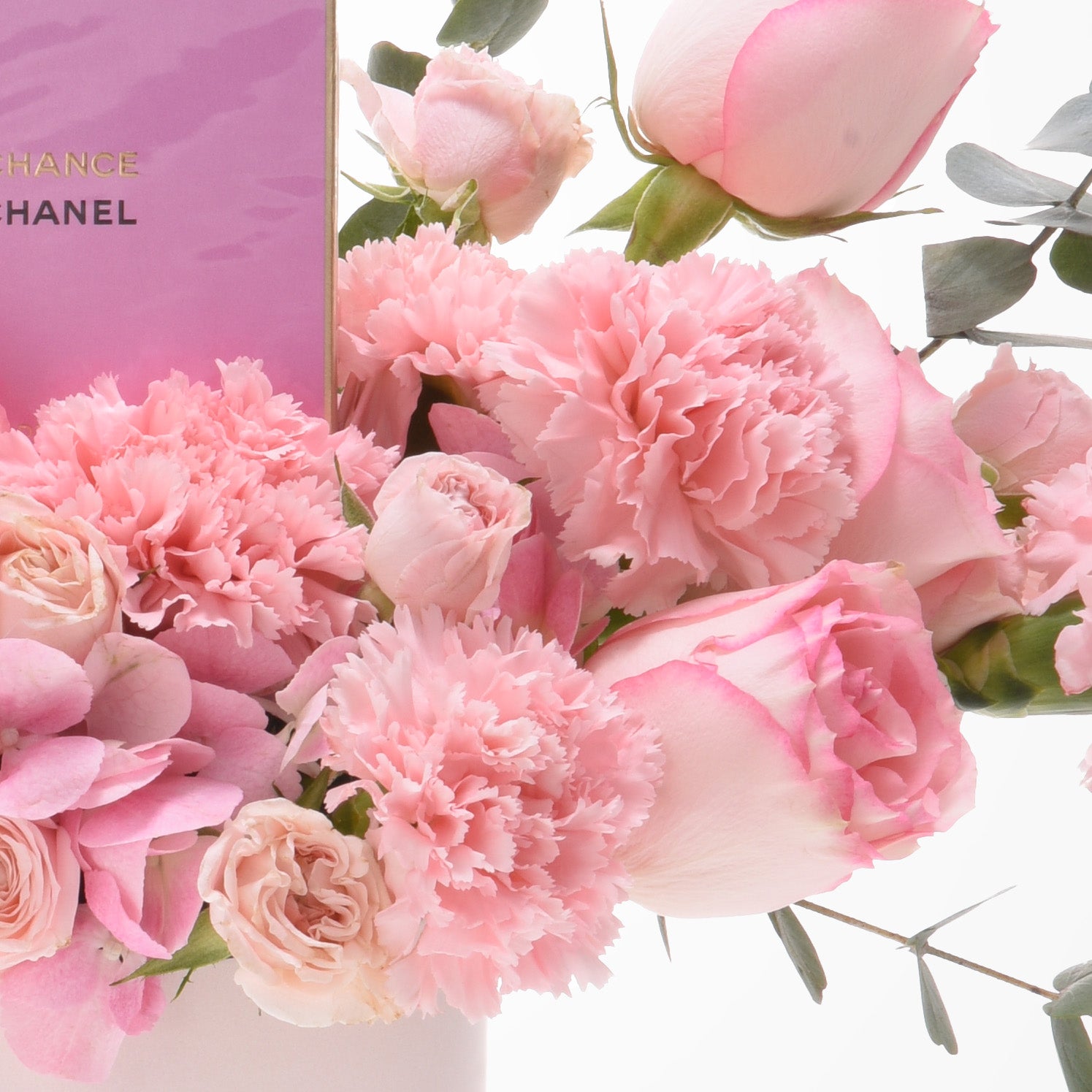 Pink roses, carnations, eucalyptus beside Chanel Chance perfume box, soft pastel luxury floral arrangement.