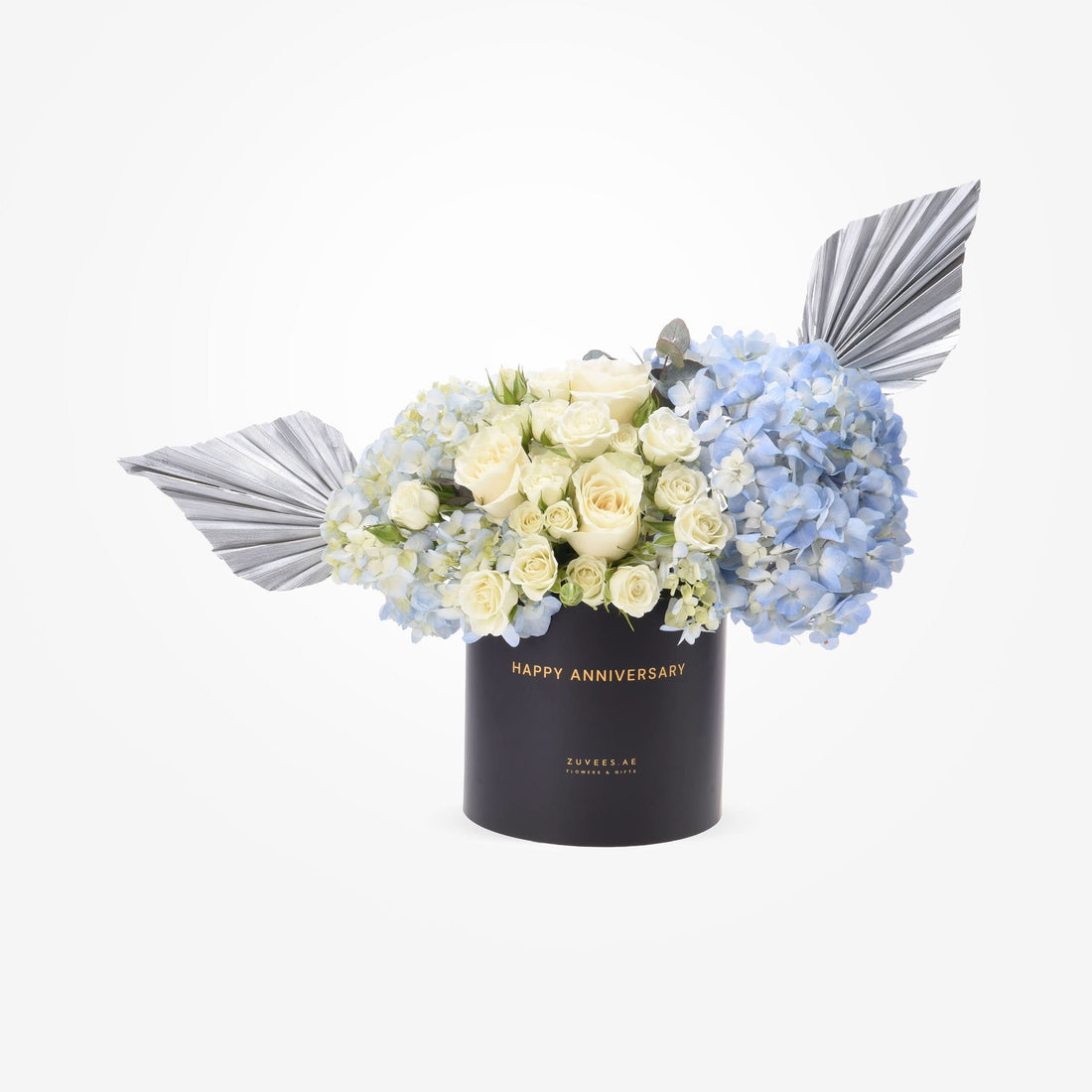 Elegant black box with "Happy Anniversary" holds creamy roses and blue hydrangeas, accented by silver fan leaves.