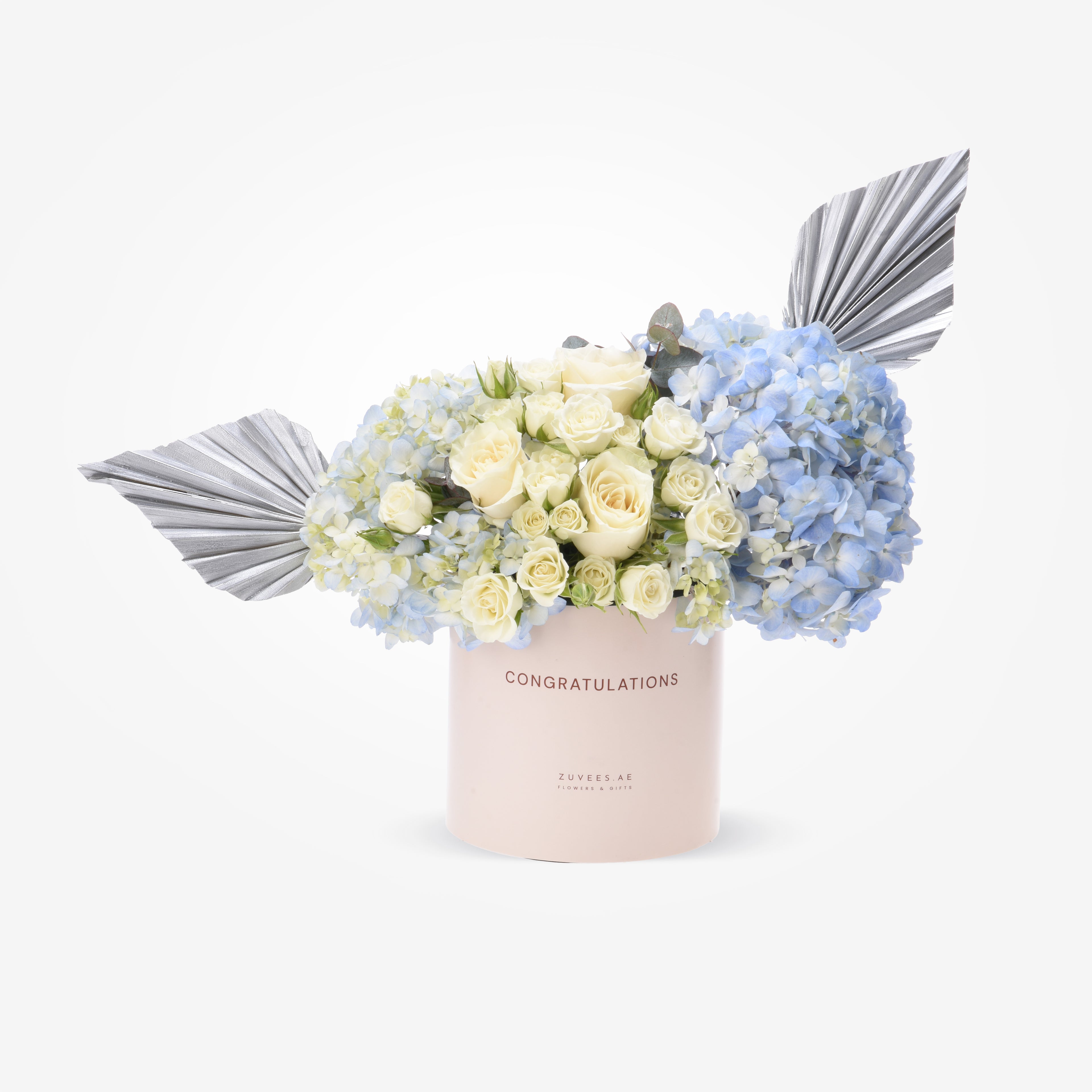 Pastel pink box with white roses and blue hydrangeas, accented by silver fan leaves.