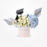 Elegant graduation bouquet with white roses, blue hydrangeas, and silver wings in a blush box.