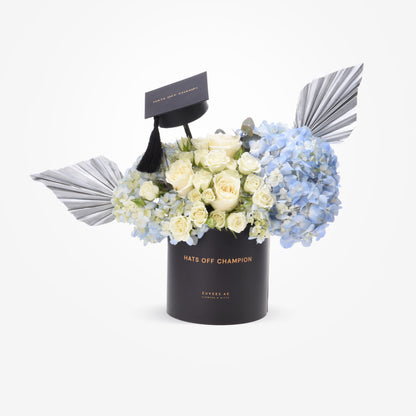 Black graduation cap atop white roses and blue hydrangeas in sleek black cylinder.