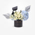 Black graduation cap atop white roses and blue hydrangeas in sleek black cylinder.