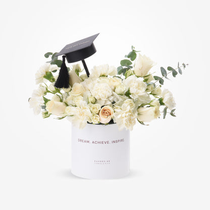 White roses and eucalyptus in sleek box topped with graduation cap.