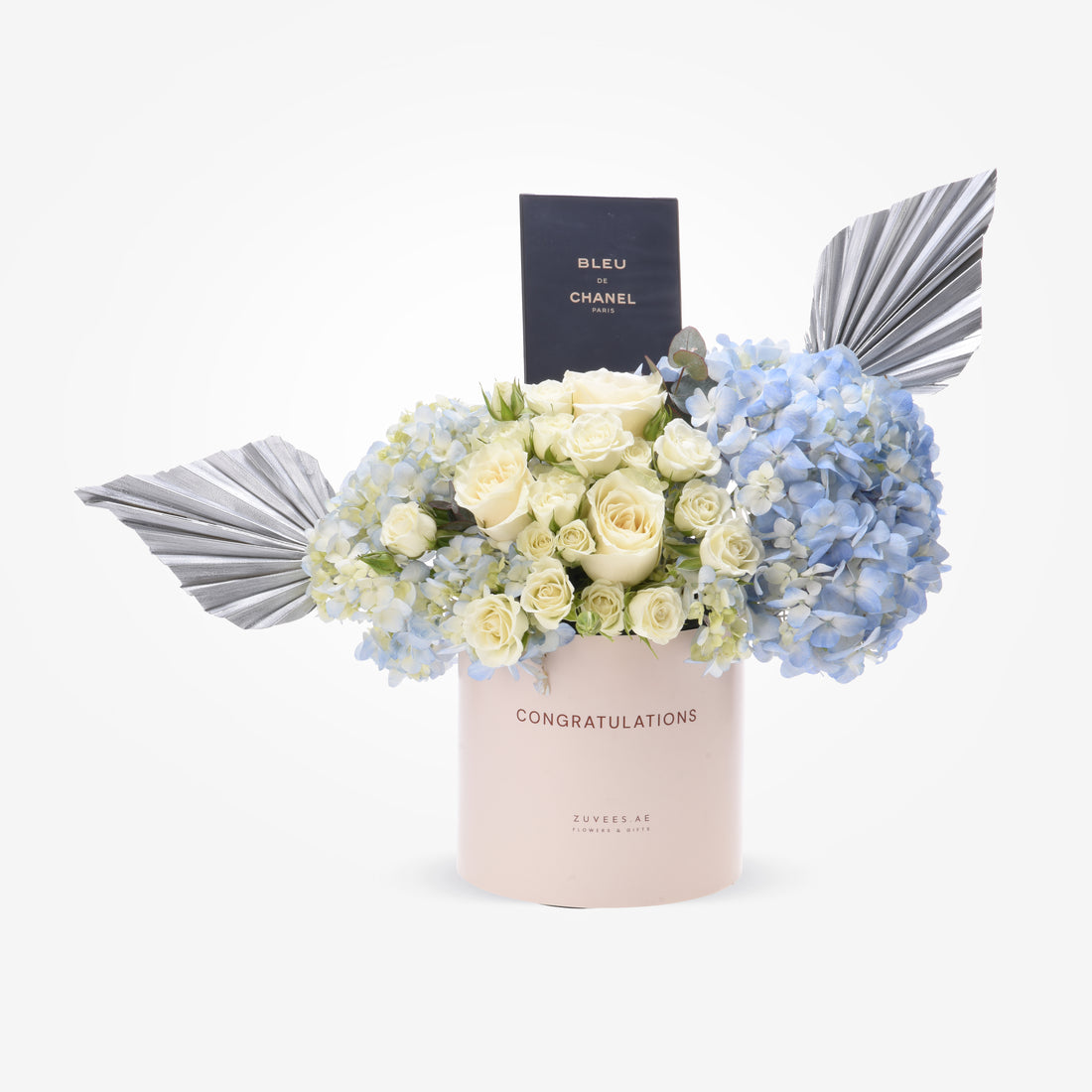 Elegant pastel pink box with "Congratulations" text, holding white roses and blue hydrangeas, accented with silver fan leaves and a Bleu de Chanel perfume bottle.