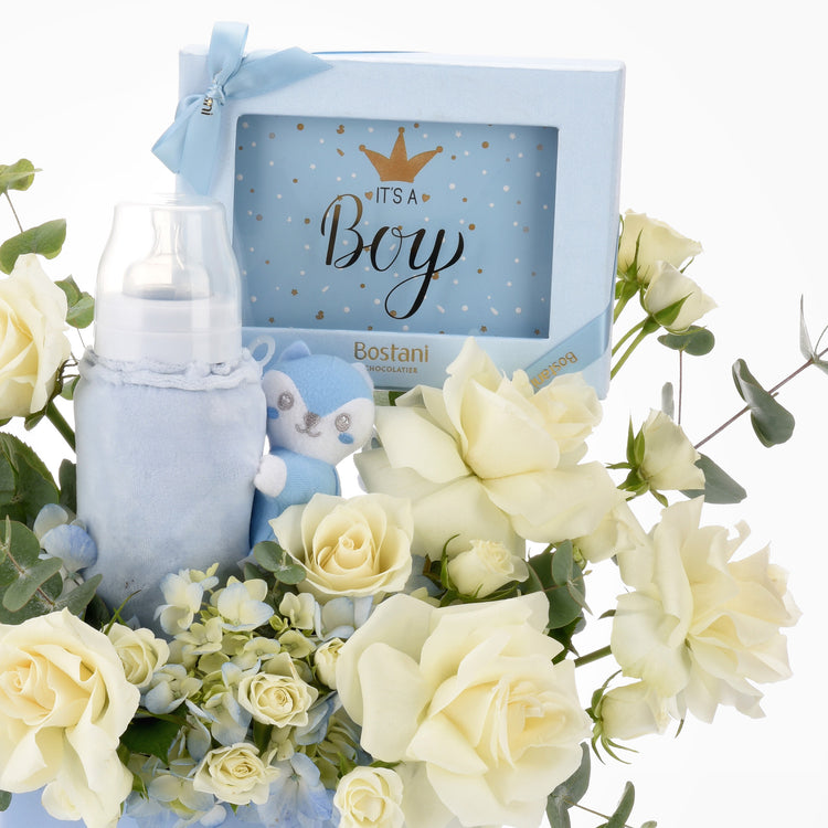Soft blue baby bottle, plush toy, and chocolate gift box nestled in creamy white roses.