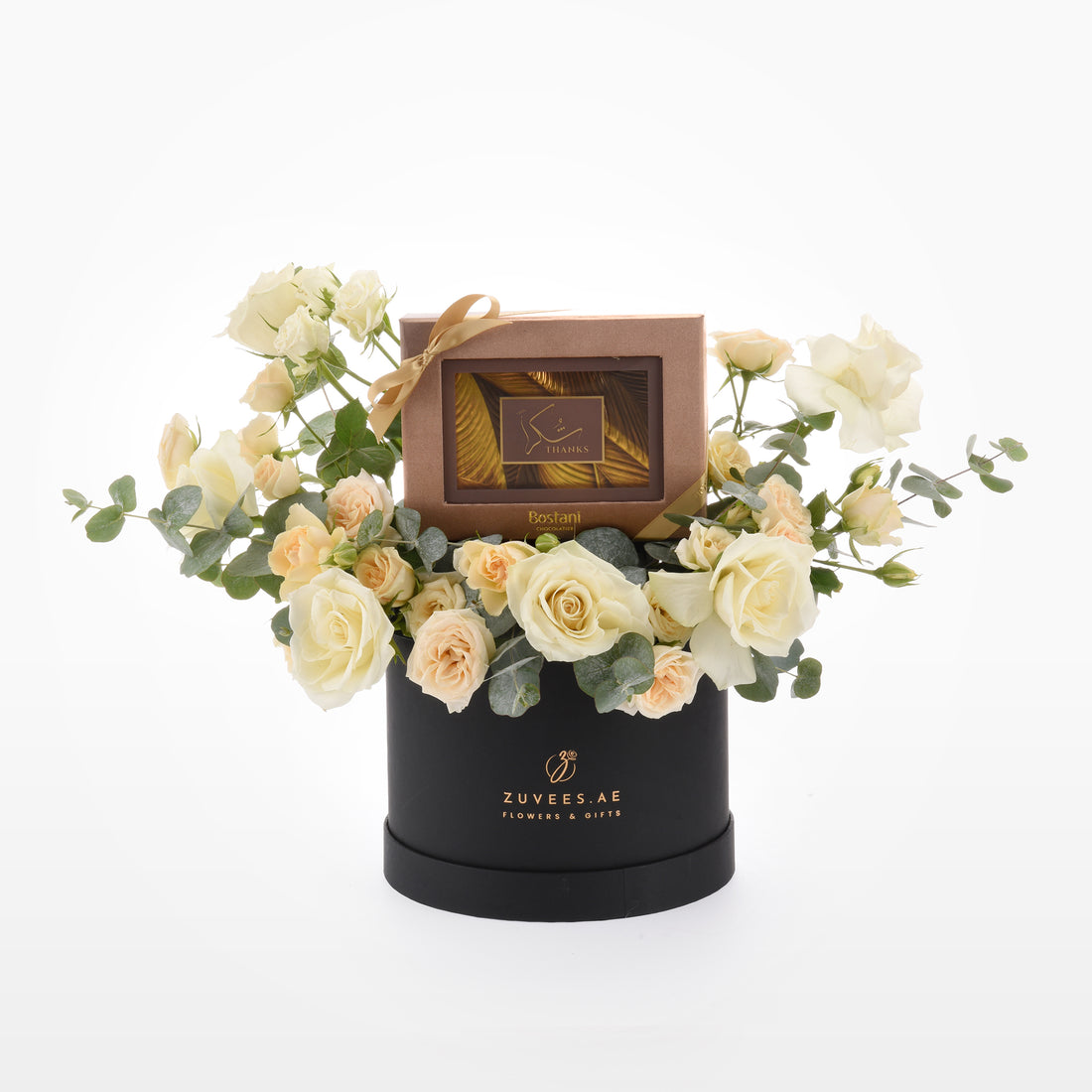 Elegant cream roses and eucalyptus surround a gold-banded chocolate box in sleek black hat.