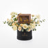 Elegant cream roses and eucalyptus surround a gold-banded chocolate box in sleek black hat.