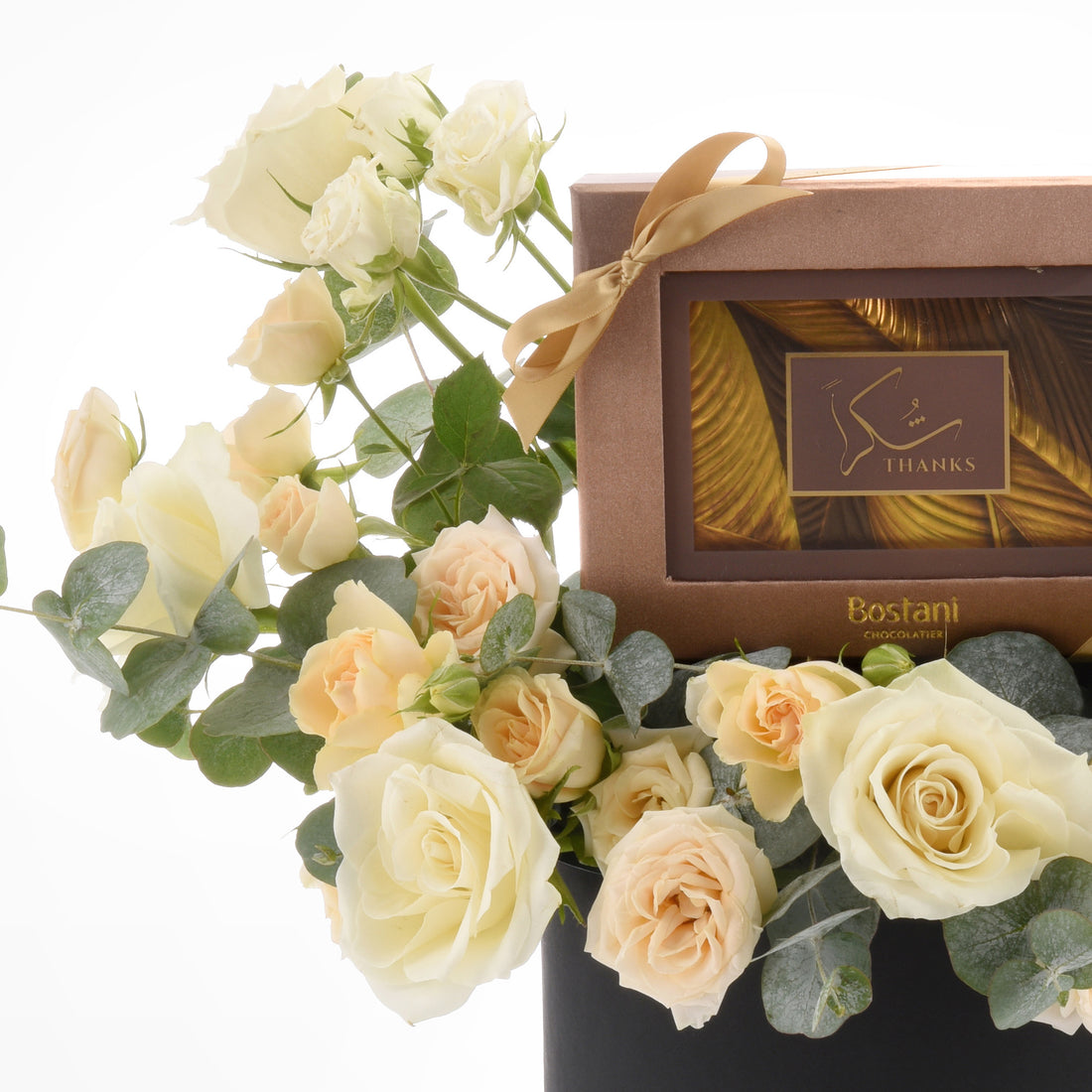 Cream roses and eucalyptus flank a gold-ribboned Bostani chocolate box.
