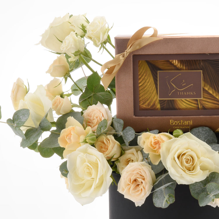 Cream roses and eucalyptus flank a gold-ribboned Bostani chocolate box.