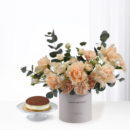 Soft peach roses, eucalyptus in grey "Happy Birthday" vase beside mini tiramisu cake.