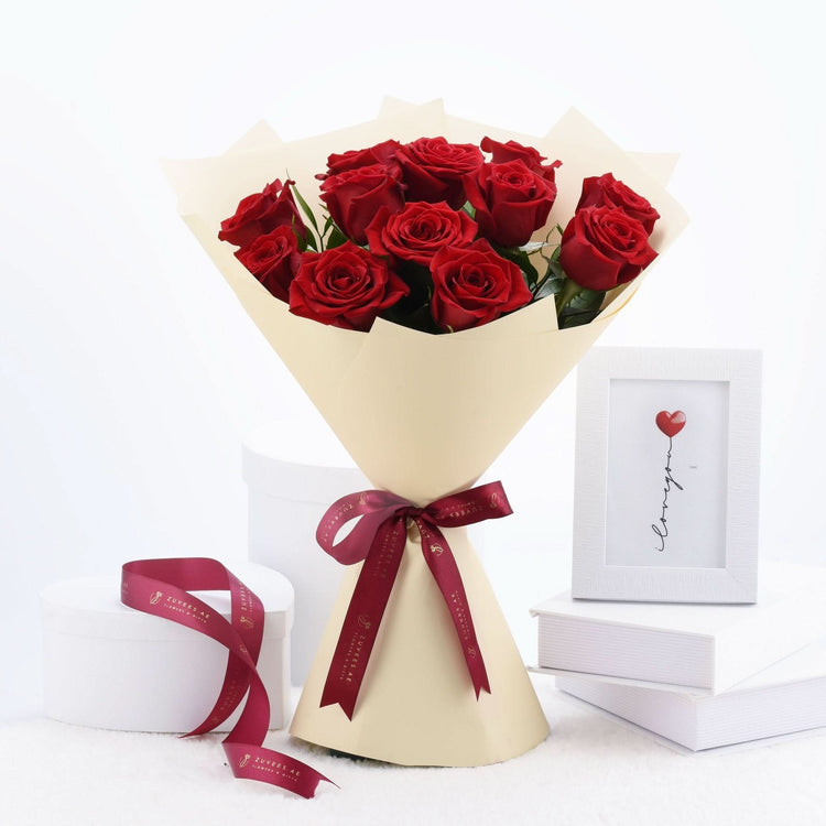 Vibrant red roses in cream paper, tied with burgundy ribbon, beside framed “I love you” art.