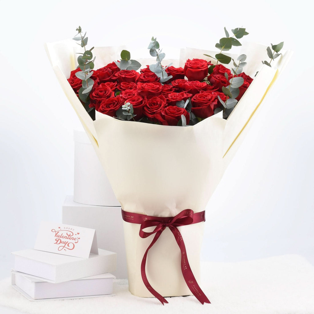 Luxurious red roses wrapped in cream paper, tied with burgundy ribbon, accented with eucalyptus, beside Valentine’s card.