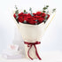 Luxurious red roses wrapped in cream paper, tied with burgundy ribbon, accented with eucalyptus, beside Valentine’s card.