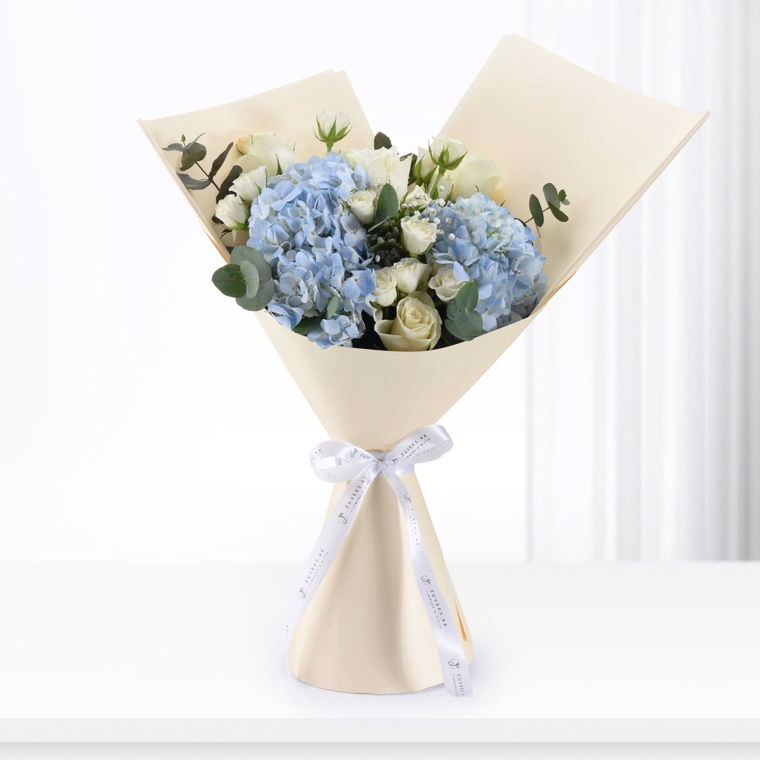 Elegant bouquet of blue hydrangeas and white roses, wrapped in cream paper with branded white ribbon.