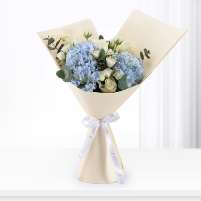Elegant bouquet of blue hydrangeas and white roses, wrapped in cream paper with branded white ribbon.