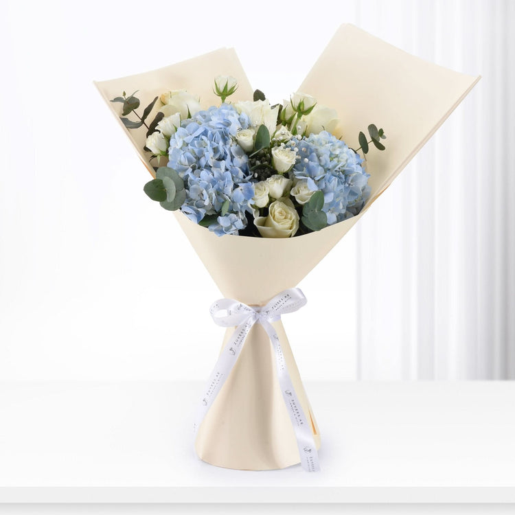 Elegant bouquet of blue hydrangeas and white roses, wrapped in cream paper with branded white ribbon.
