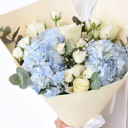 Elegant bouquet of white roses and blue hydrangeas, wrapped in cream paper with eucalyptus.