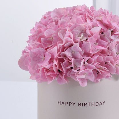 Soft pink hydrangeas in minimalist white vase with "HAPPY BIRTHDAY" text.
