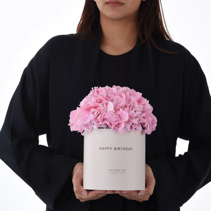 Pink hydrangeas burst from a white "Happy Birthday" box held by a person in black.