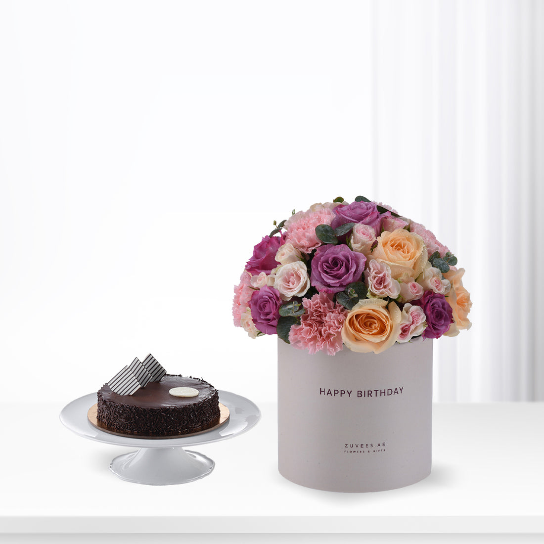Purple, pink, and peach roses in grey "Happy Birthday" cylinder beside chocolate cake.