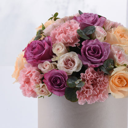 Soft pink, purple, and peach roses with carnations and dewdrops in a silver vase.