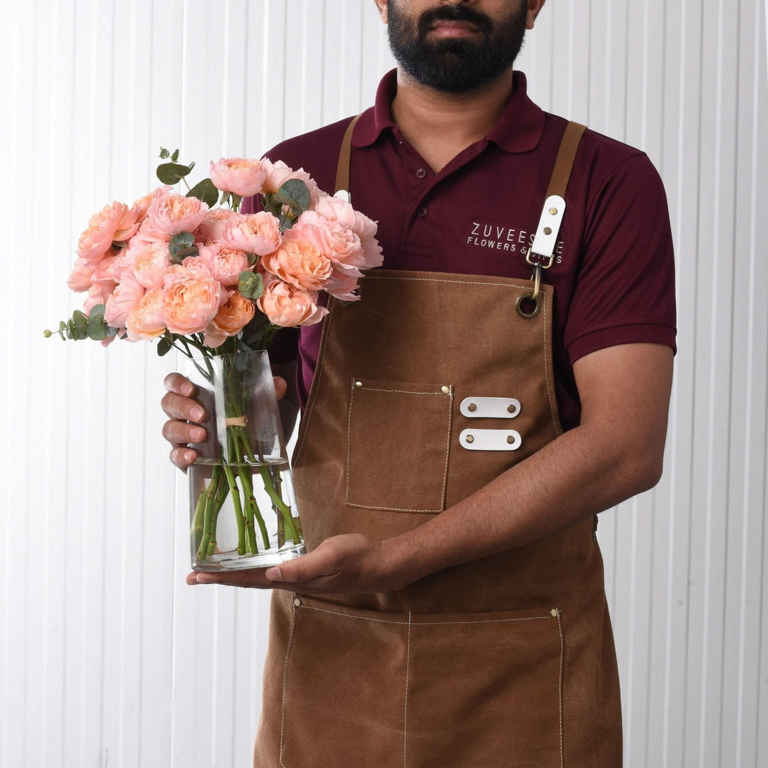 Brown apron-wearing florist holds glass vase of peach roses against white corrugated backdrop.