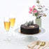 Rich chocolate cake with striped chocolate garnish, champagne flutes, and pink floral arrangement.