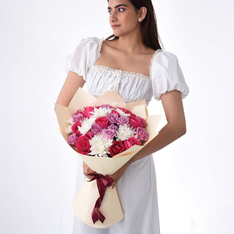 Woman holds vibrant pink, purple, white rose bouquet wrapped in cream paper with burgundy ribbon.