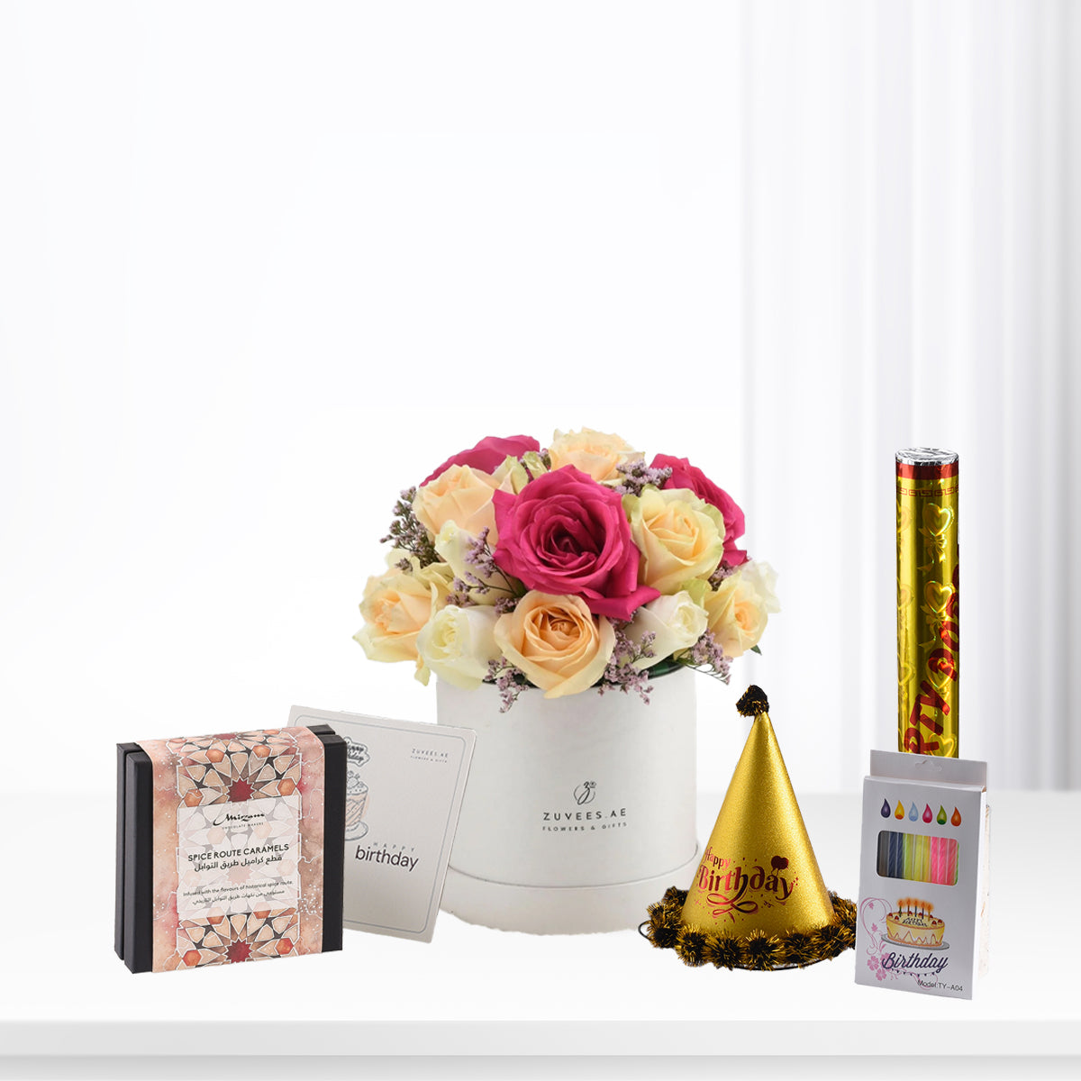 Vibrant pink and cream roses in white box, gold party hat, caramel gift, and birthday candles.