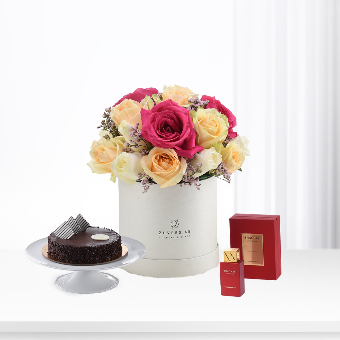 Vibrant pink, yellow roses in white box beside chocolate cake and red perfume.