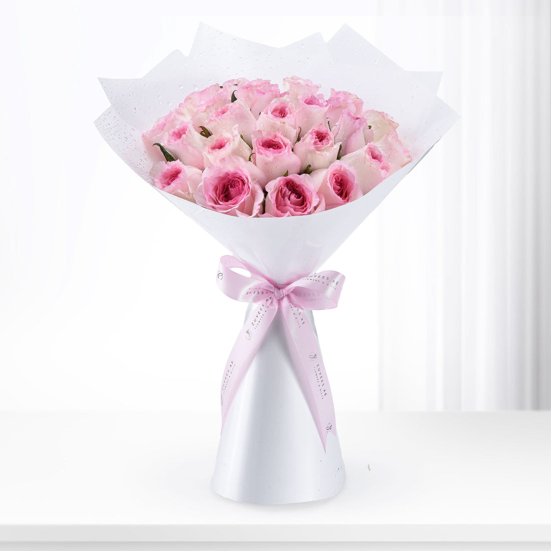 Pink roses in white wrap with dewdrops, tied with branded ribbon, elegant gift bouquet.