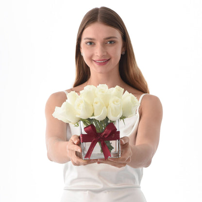 Woman holds white roses in clear vase with burgundy ribbon, smiling against pure white background.
