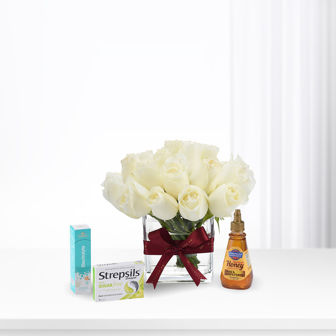 White roses in glass vase with burgundy bow, beside Strepsils, honey, and electrolyte.