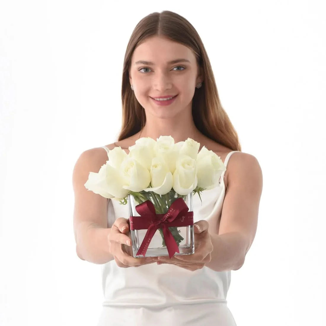 Woman holds white roses in clear vase with red ribbon, smiling against pure white background.