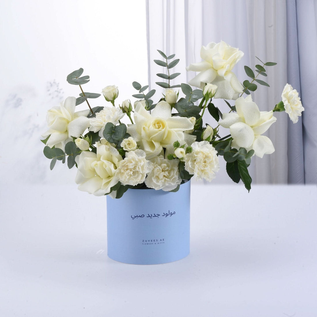 White roses and carnations in light blue cylinder vase with Arabic greeting.
