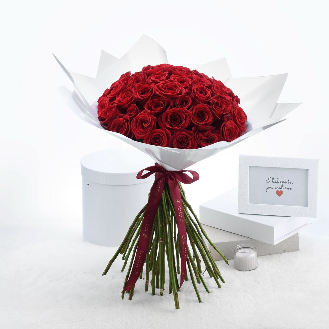 Vibrant red roses in white paper, tied with burgundy ribbon, beside framed message and candle.