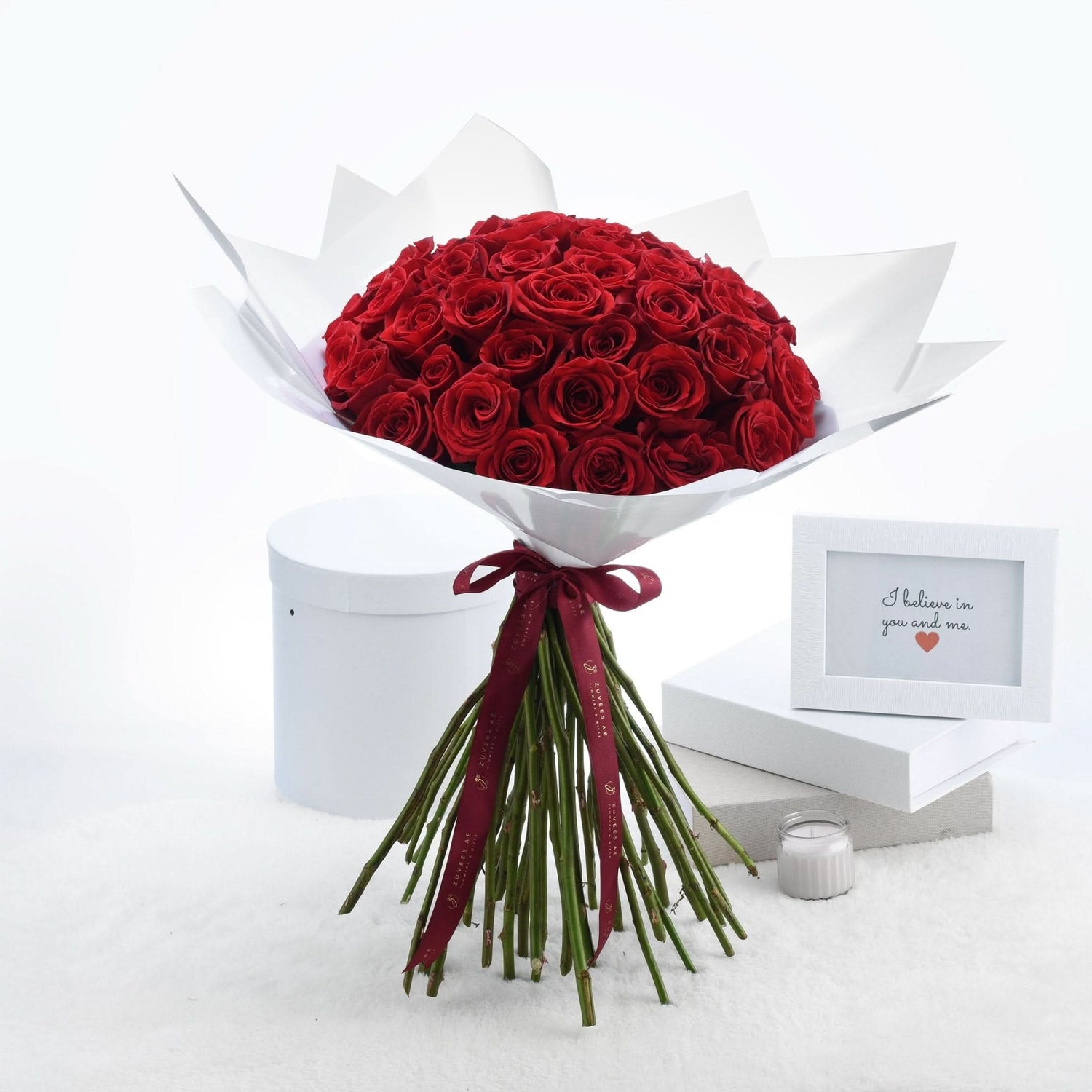 Vibrant red roses in white paper, tied with burgundy ribbon, beside framed message and candle.