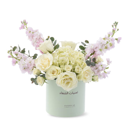 Soft pastel floral arrangement in mint container with Arabic branding.