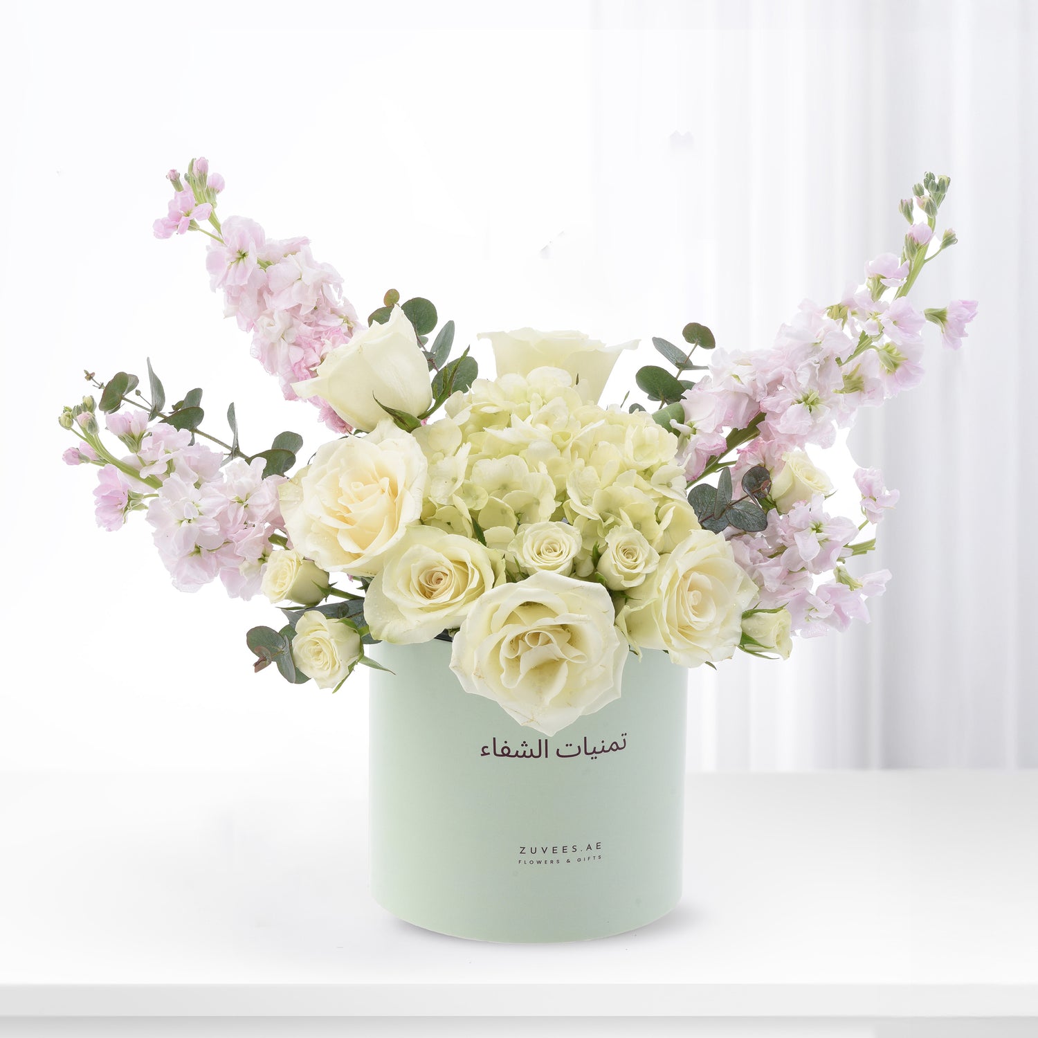Pastel green cylinder vase holds creamy white roses and soft pink blooms against bright white background.