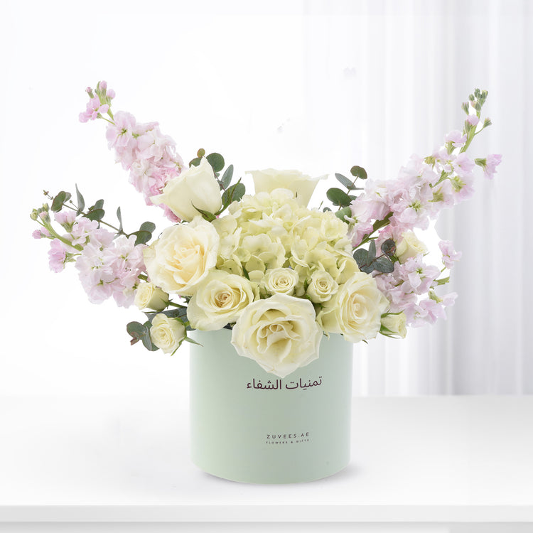 Pastel green cylinder vase holds creamy white roses and soft pink blooms against bright white background.