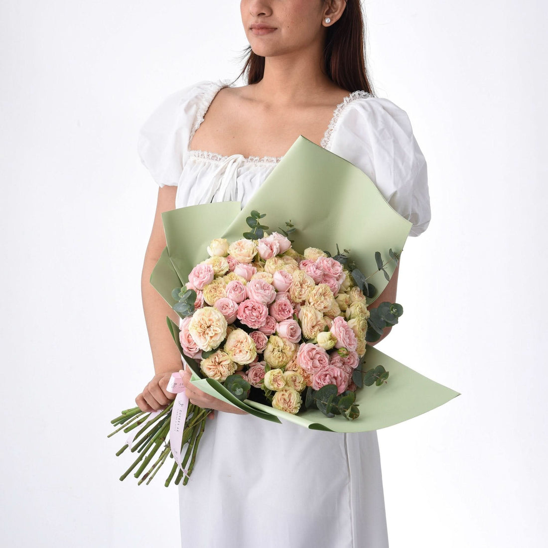 Soft pink and cream roses wrapped in sage green paper, held by a woman in white.