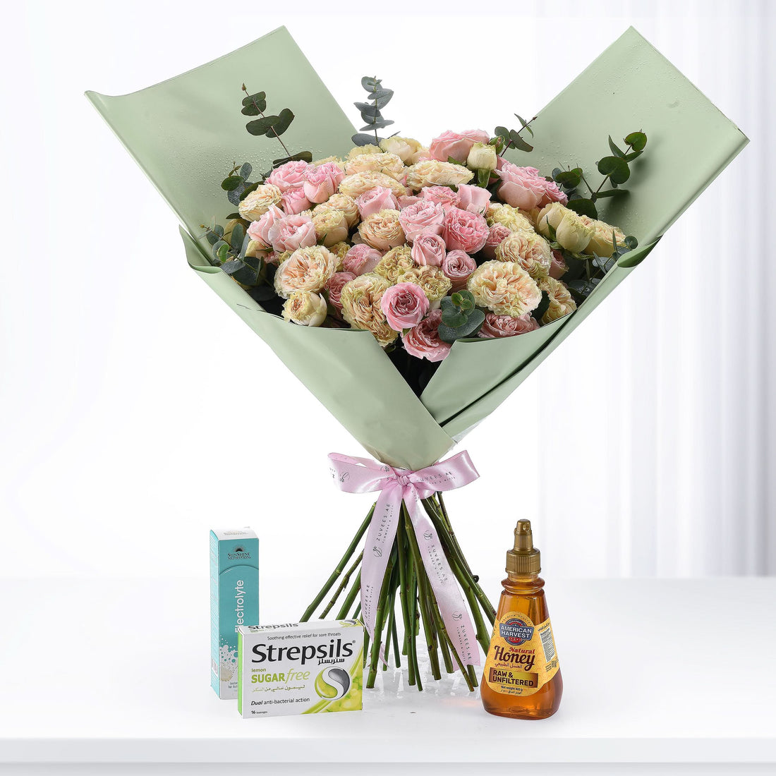 Soft pink and cream roses wrapped in sage green paper, tied with pink ribbon, beside Strepsils and honey.