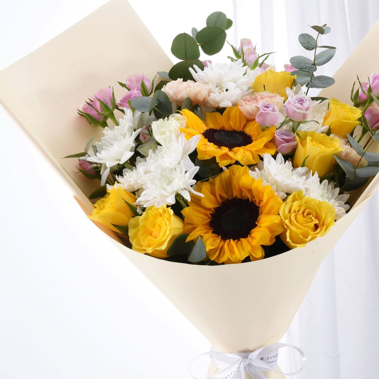 Vibrant sunflowers, roses, and chrysanthemums in creamy wrap with elegant white bow.