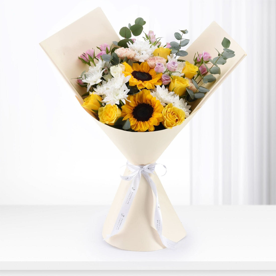 Vibrant sunflowers, roses, and chrysanthemums in creamy wrap with elegant white ribbon.