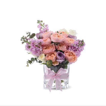 Pastel pink, peach, and lavender roses with baby’s breath in clear vase, pink ribbon bow.