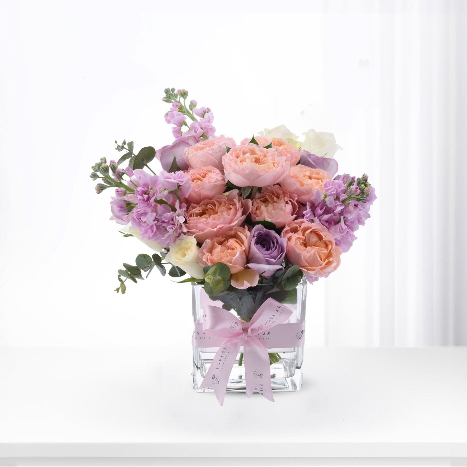 Pastel pink, peach, and lavender roses with eucalyptus in clear glass vase, tied with branded pink ribbon.