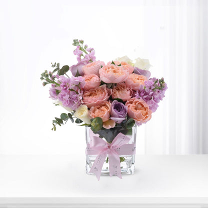 Pastel pink, peach, and lavender roses with eucalyptus in clear glass vase, tied with branded pink ribbon.
