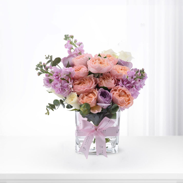 Pastel pink, peach, and lavender roses with eucalyptus in clear glass vase, tied with branded pink ribbon.