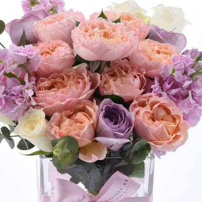Pastel pink, lavender, and cream roses elegantly arranged in clear glass vase with pink ribbon.
