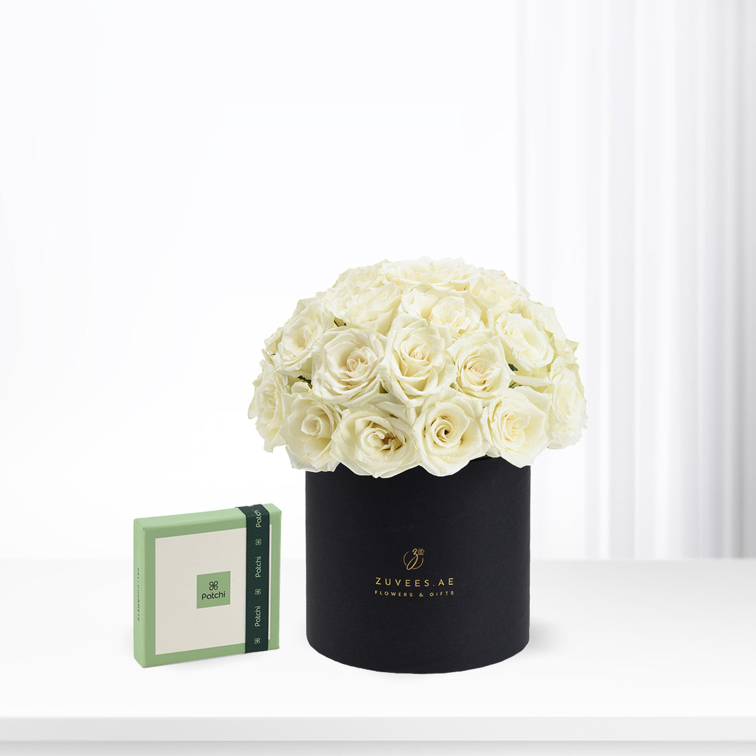 Elegant ivory roses in matte black hat box beside green Patchi gift box.